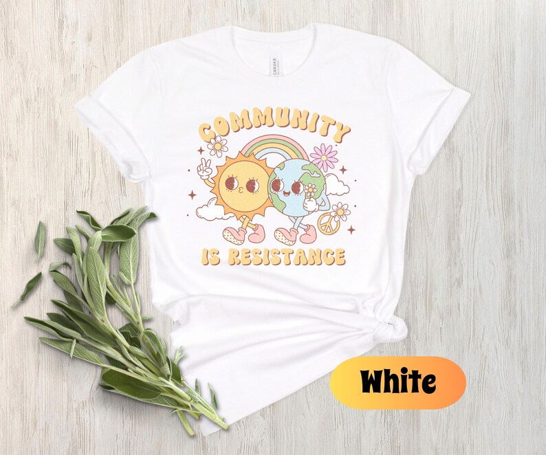 May include: White t-shirt with a graphic of a sun and earth holding hands with the text "Community is Resistance".