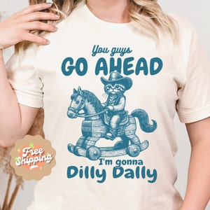 May include: A white t-shirt with a blue graphic of a cat riding a rocking horse. The text "You guys go ahead I'm gonna dilly dally" is printed on the shirt.