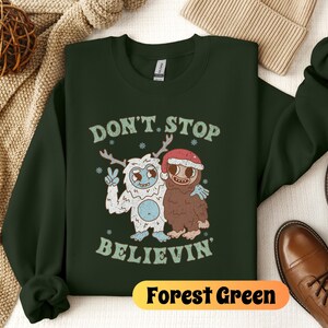 May include: Forest green sweatshirt with a white yeti and a brown yeti wearing a red Santa hat. The text "Don't Stop Believin'" is printed on the sweatshirt.