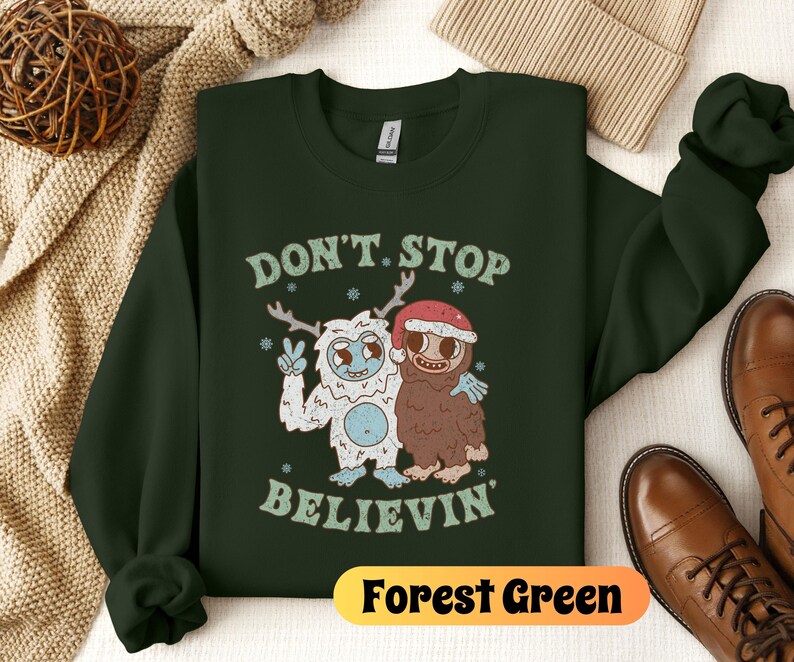 May include: Forest green sweatshirt with a white yeti and a brown yeti wearing a red Santa hat. The text "Don't Stop Believin'" is printed on the sweatshirt.