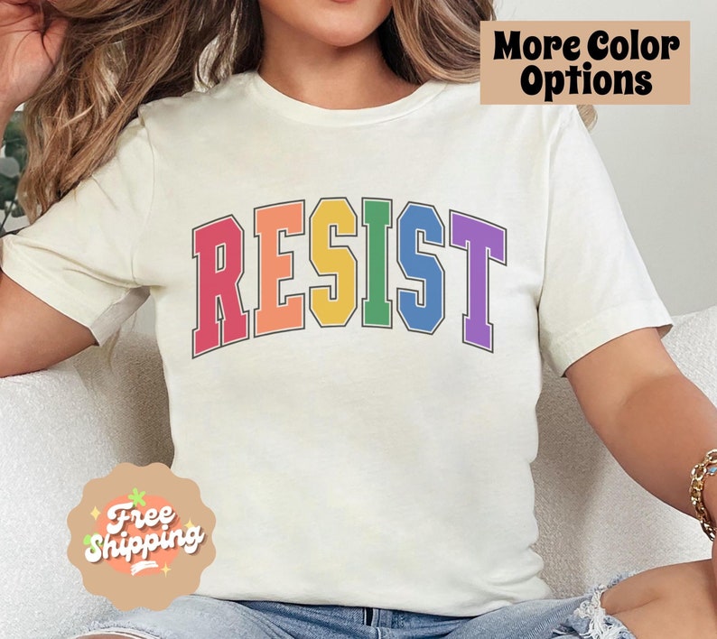 May include: A white t-shirt with the word "RESIST" printed in rainbow colors. The letters are outlined in black and have a varsity style font.