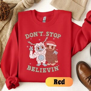 May include: Red Christmas sweatshirt with a white yeti and a brown monster holding hands. The text on the sweatshirt says "Don't Stop Believin'"