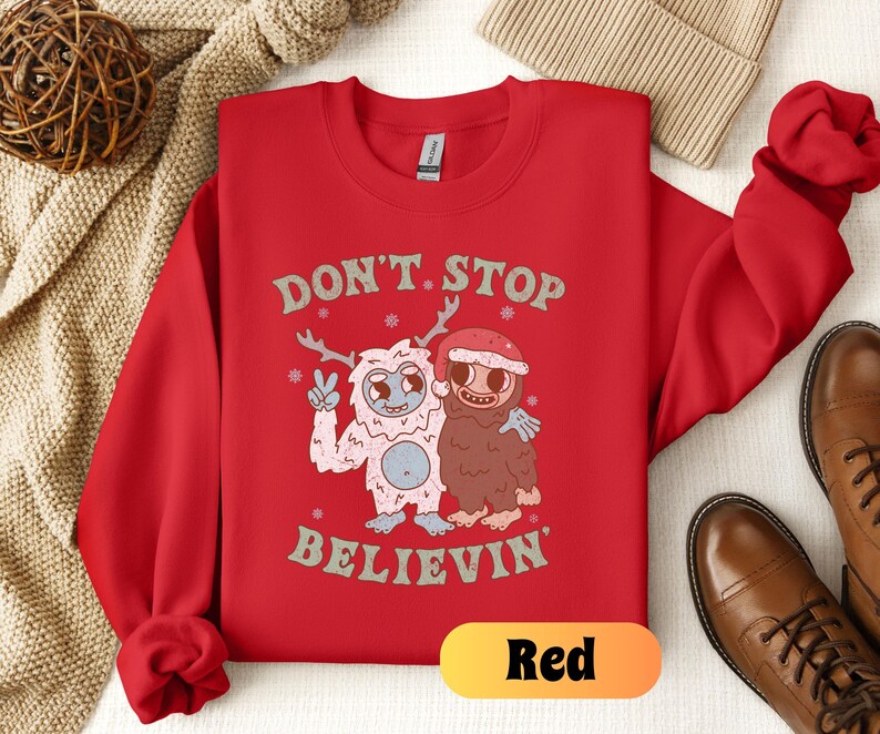 May include: Red Christmas sweatshirt with a white yeti and a brown monster holding hands. The text on the sweatshirt says "Don't Stop Believin'"