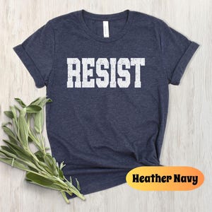 May include: A navy blue t-shirt with the word "RESIST" printed in bold, white, block letters. The shirt is a short-sleeved crew neck. The words "Heather Navy" are printed on an orange oval.