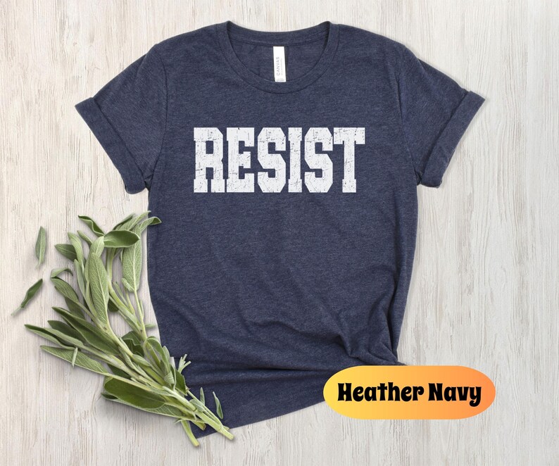 May include: A navy blue t-shirt with the word "RESIST" printed in bold, white, block letters. The shirt is a short-sleeved crew neck. The words "Heather Navy" are printed on an orange oval.