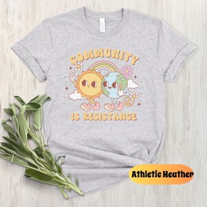 May include: Gray t-shirt with a graphic of a sun and earth holding hands with the text "Community is Resistance".