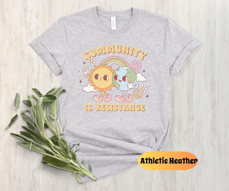 May include: Gray t-shirt with a graphic of a sun and earth holding hands with the text "Community is Resistance".
