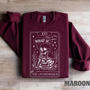 Puede incluir: Sudadera granate con un gr&aacute;fico blanco de un esqueleto sosteniendo una taza, con el texto "WHAT IF..." y "THE OVERTHINKER". La sudadera tiene cuello redondo y mangas largas.
