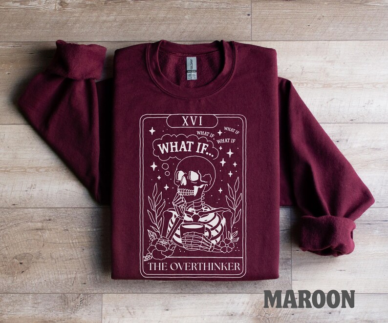 Puede incluir: Sudadera granate con un gr&aacute;fico blanco de un esqueleto sosteniendo una taza, con el texto "WHAT IF..." y "THE OVERTHINKER". La sudadera tiene cuello redondo y mangas largas.