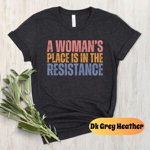 May include: Dark grey heather t-shirt with the text "A Woman's Place Is In The Resistance" in pink, gold, and blue. The shirt is displayed on a light wood surface with a bundle of green sage.