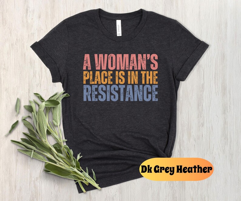 May include: Dark grey heather t-shirt with the text "A Woman's Place Is In The Resistance" in pink, gold, and blue. The shirt is displayed on a light wood surface with a bundle of green sage.