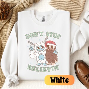 May include: White crewneck sweatshirt with a Christmas-themed graphic featuring a yeti and a sloth. The graphic says "Don't Stop Believin".