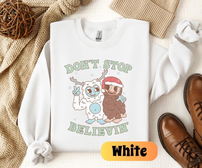 May include: White crewneck sweatshirt with a Christmas-themed graphic featuring a yeti and a sloth. The graphic says "Don't Stop Believin".