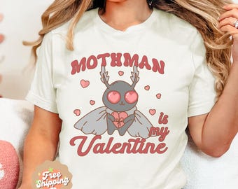May include: White t-shirt with a pink and grey graphic of a mothman holding a heart. The text "Mothman is my Valentine" is printed in pink on the shirt.