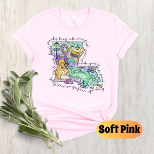 Op de afbeelding: Een zacht roze T-shirt met een kleurrijke illustratie van de staat Louisiana. De illustratie bevat Mardi Gras-parels, een koningentaart, een fleur-de-lis en een paradewagen. De tekst "laissez les bon temps rouler" staat rond de omtrek van de staat.