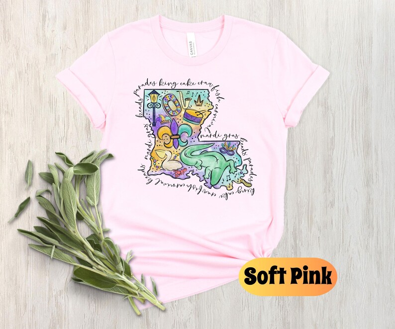 Op de afbeelding: Een zacht roze T-shirt met een kleurrijke illustratie van de staat Louisiana. De illustratie bevat Mardi Gras-parels, een koningentaart, een fleur-de-lis en een paradewagen. De tekst "laissez les bon temps rouler" staat rond de omtrek van de staat.