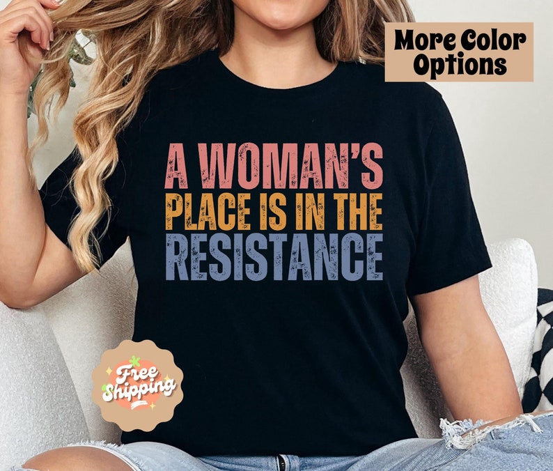 May include: Black t-shirt with the text "A Woman's Place Is In The Resistance" in pink, yellow, and blue. The shirt has a vintage, distressed look. Additional text in the image says "More Color Options" and "Free Shipping".