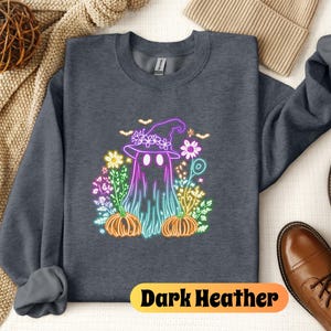 K&ouml;nnte beinhalten: Dunkelgrauer Sweatshirt mit einem Neon-Regenbogen-Motiv eines Geistes, der einen Hexenhut tr&auml;gt und von Blumen und K&uuml;rbissen umgeben ist. Der Text "Dark Heather" ist unten auf dem Sweatshirt aufgedruckt.