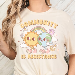 May include: A cream colored t-shirt with a graphic of a smiling sun and earth holding hands. The text "COMMUNITY IS RESISTANCE" is printed in a retro font.