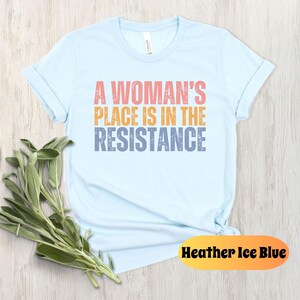 May include: Light blue t-shirt with the text "A Woman's Place Is In The Resistance" in coral, orange, and blue. The shirt is made of soft material and has rolled-up sleeves. A sprig of green leaves is placed near the shirt. The color is "Heather Ice Blue."