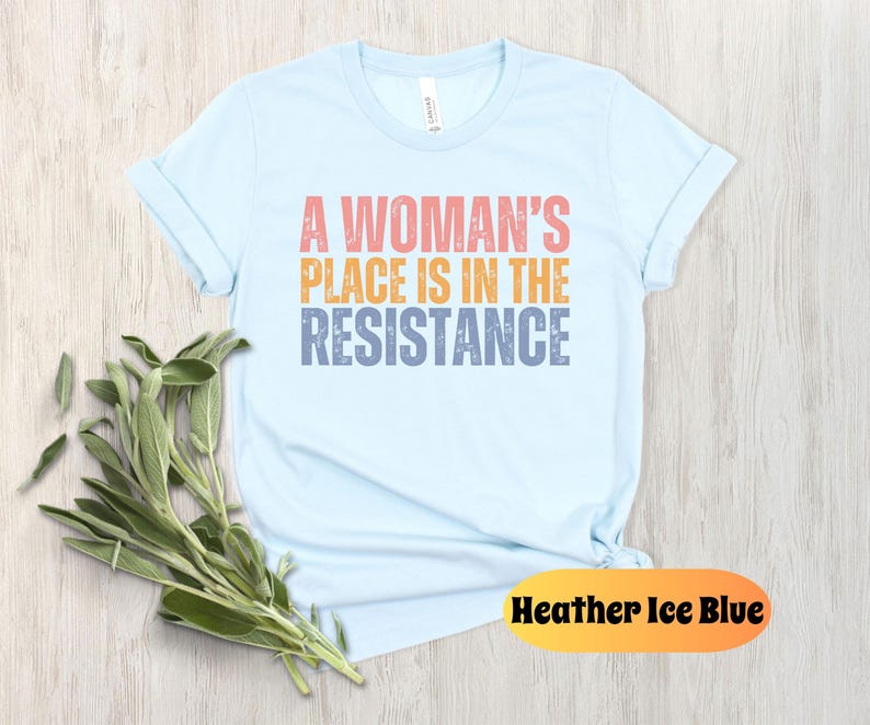 May include: Light blue t-shirt with the text "A Woman's Place Is In The Resistance" in coral, orange, and blue. The shirt is made of soft material and has rolled-up sleeves. A sprig of green leaves is placed near the shirt. The color is "Heather Ice Blue."