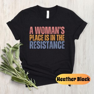 May include: A black t-shirt with the text "A Woman's Place Is In The Resistance" in pink, yellow, and blue. The shirt is made of a soft material and is a classic crew neck style. The words "Heather Black" are in a yellow oval.