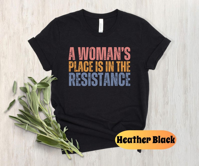 May include: A black t-shirt with the text "A Woman's Place Is In The Resistance" in pink, yellow, and blue. The shirt is made of a soft material and is a classic crew neck style. The words "Heather Black" are in a yellow oval.