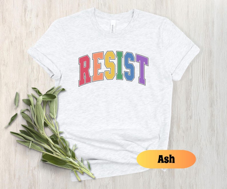 May include: A light grey t-shirt with the word "RESIST" printed in rainbow colors. The word is in a varsity style font with a black outline. The t-shirt is available in the color Ash.