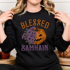 以下が含まれることがあります： 紫色の文字で「Blessed Samhain」と書かれた黒いスウェットシャツ。シャツの中央には、紫の花がついたカボチャの漫画が描かれている。
