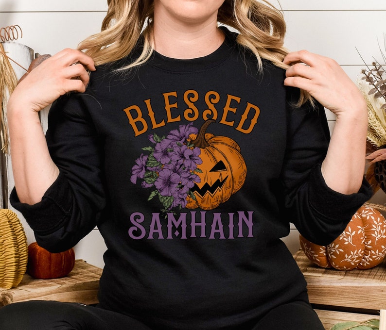 以下が含まれることがあります： 紫色の文字で「Blessed Samhain」と書かれた黒いスウェットシャツ。シャツの中央には、紫の花がついたカボチャの漫画が描かれている。