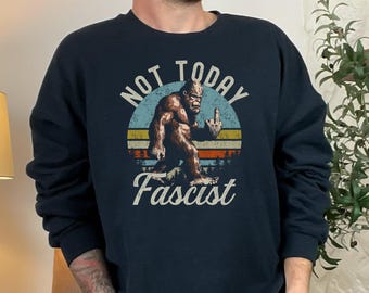 May include: A dark blue sweatshirt featuring a retro-style graphic of a bigfoot giving the middle finger. The graphic is surrounded by a red, white and blue colour scheme and the text "Not Today Fascist".
