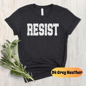 May include: Dark grey heather t-shirt with the word "RESIST" printed in large, distressed white block letters. The shirt has a crew neck and rolled-up sleeves. A sprig of green leaves is in the lower left corner. The text "Dk Grey Heather" is in an orange oval.