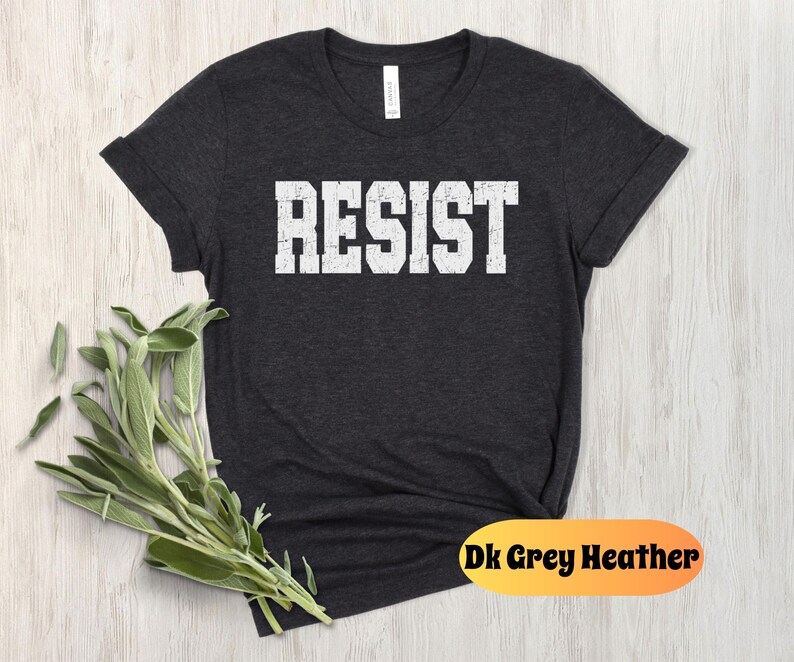 May include: Dark grey heather t-shirt with the word "RESIST" printed in large, distressed white block letters. The shirt has a crew neck and rolled-up sleeves. A sprig of green leaves is in the lower left corner. The text "Dk Grey Heather" is in an orange oval.