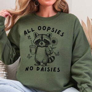 May include: Olive green sweatshirt with a raccoon graphic holding a drink and a sunflower. The text "ALL OOPSIES NO DAISIES" is printed above and below the raccoon illustration. Butterflies are also included in the design.
