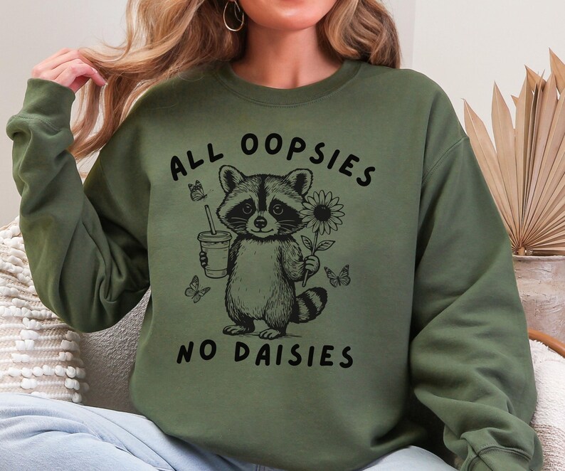 May include: Olive green sweatshirt with a raccoon graphic holding a drink and a sunflower. The text "ALL OOPSIES NO DAISIES" is printed above and below the raccoon illustration. Butterflies are also included in the design.