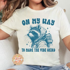 May include: White t-shirt with a blue graphic of a raccoon wearing a hoodie and backpack. The text "On My Way To Make The Vibe Weird" is printed in blue below the raccoon.