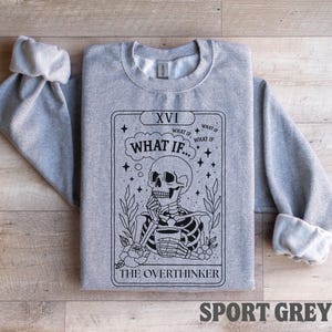 Puede incluir: Una sudadera gris claro con mangas enrolladas. La parte delantera presenta un dise&ntilde;o de carta del tarot en blanco y negro con un esqueleto, las palabras "WHAT IF..." y "THE OVERTHINKER". El texto "SPORT GREY" est&aacute; en la parte inferior.
