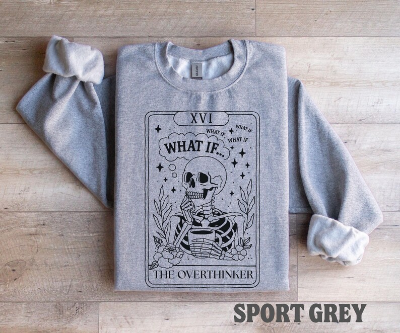 Puede incluir: Una sudadera gris claro con mangas enrolladas. La parte delantera presenta un dise&ntilde;o de carta del tarot en blanco y negro con un esqueleto, las palabras "WHAT IF..." y "THE OVERTHINKER". El texto "SPORT GREY" est&aacute; en la parte inferior.