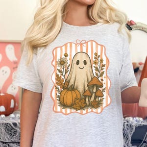 May include: Light gray t-shirt featuring a vintage-style Halloween graphic. The design includes a smiling ghost, pumpkin, mushrooms, and fall foliage within a peach-colored frame with vertical stripes. A cute and spooky design for the Halloween season.