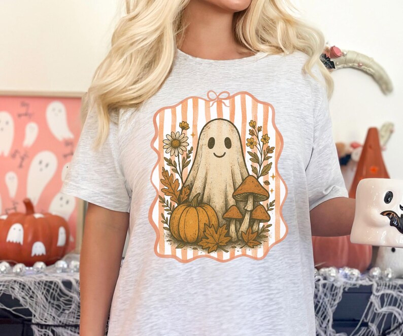 May include: Light gray t-shirt featuring a vintage-style Halloween graphic. The design includes a smiling ghost, pumpkin, mushrooms, and fall foliage within a peach-colored frame with vertical stripes. A cute and spooky design for the Halloween season.