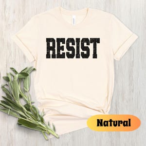 May include: A cream-coloured t-shirt featuring the word "RESIST" in distressed black block letters. The shirt has a crew neck and short sleeves. A sprig of green herbs is in the lower left corner. The word "Natural" is in a yellow and orange oval.