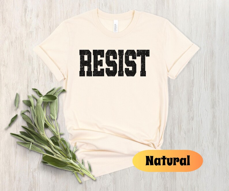May include: A cream-coloured t-shirt featuring the word "RESIST" in distressed black block letters. The shirt has a crew neck and short sleeves. A sprig of green herbs is in the lower left corner. The word "Natural" is in a yellow and orange oval.