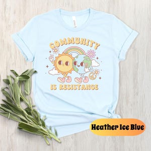 May include: Light blue t-shirt with a cartoon sun and earth holding hands. The text "Community is Resistance" is printed on the shirt.