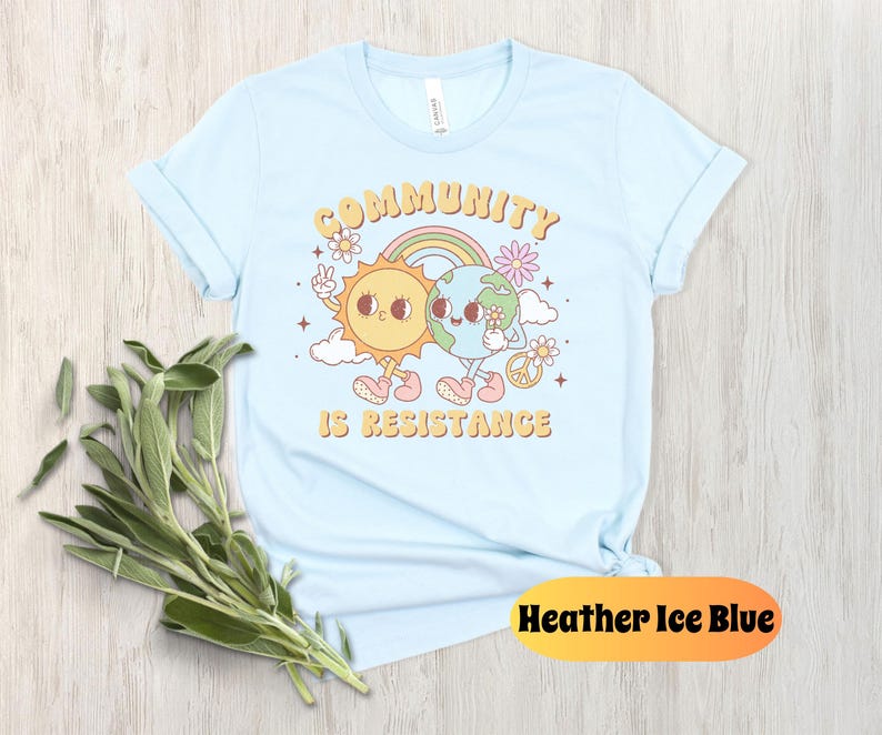 May include: Light blue t-shirt with a cartoon sun and earth holding hands. The text "Community is Resistance" is printed on the shirt.