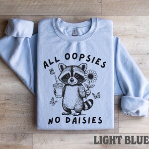 May include: Light blue sweatshirt with the text "ALL OOPSIES NO DAISIES" and a raccoon graphic holding a drink and a flower. The graphic is black and white. The words "LIGHT BLUE" are at the bottom.