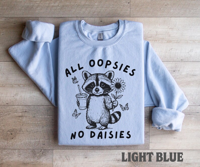 May include: Light blue sweatshirt with the text "ALL OOPSIES NO DAISIES" and a raccoon graphic holding a drink and a flower. The graphic is black and white. The words "LIGHT BLUE" are at the bottom.