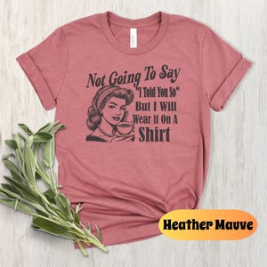 May include: Heather mauve t-shirt with a retro-style illustration of a woman drinking from a teacup. The text on the shirt reads "Not Going To Say "I Told You So" But I Will Wear It On A Shirt"