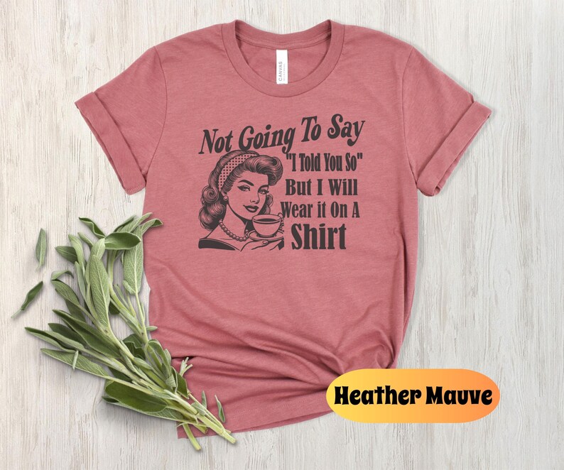 May include: Heather mauve t-shirt with a retro-style illustration of a woman drinking from a teacup. The text on the shirt reads "Not Going To Say "I Told You So" But I Will Wear It On A Shirt"