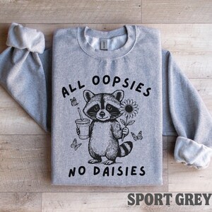 May include: A gray sweatshirt with the text "ALL OOPSIES NO DAISIES" and a raccoon graphic holding a drink and a flower. The sweatshirt is sport grey.