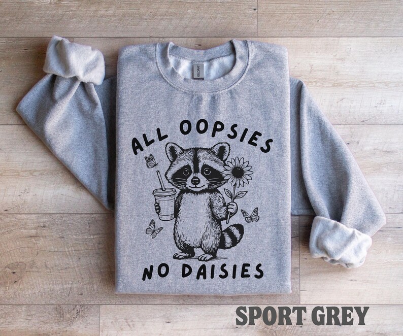 May include: A gray sweatshirt with the text "ALL OOPSIES NO DAISIES" and a raccoon graphic holding a drink and a flower. The sweatshirt is sport grey.